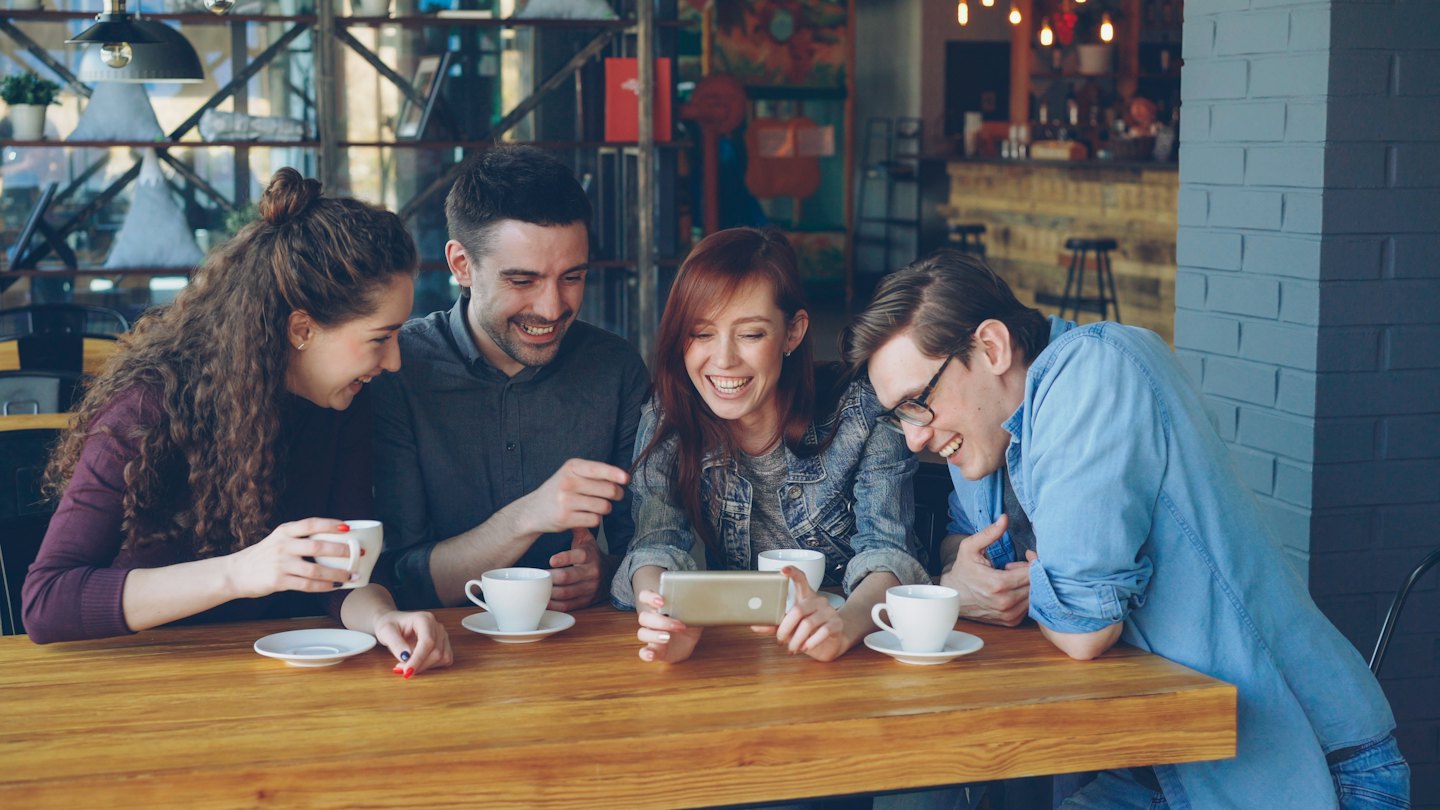 Quatre amis rient en regardant un smartphone à la table d'un café.